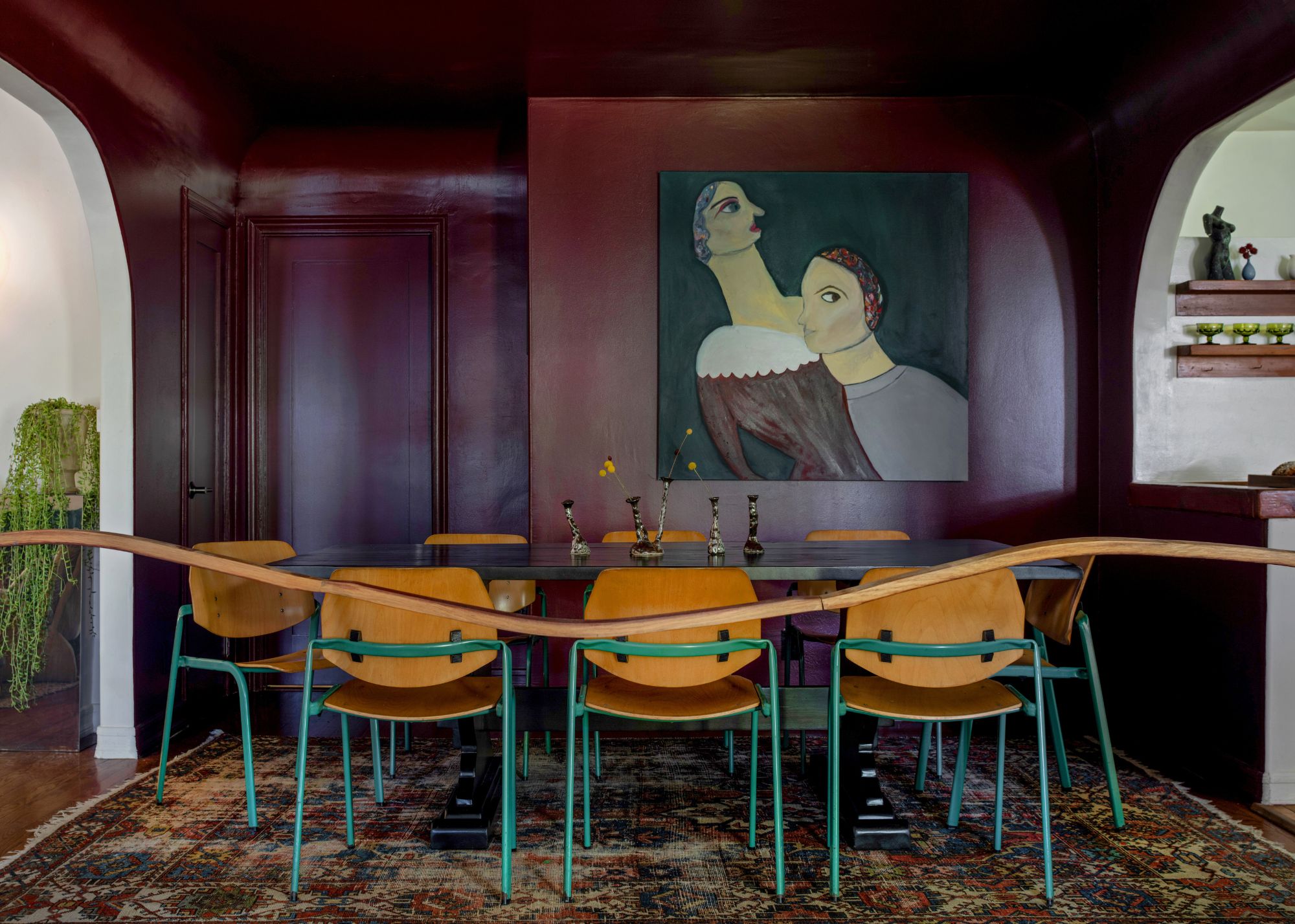 A moody dining room with dark red walls, a patterned rug, and a dining table with wooden chairs and artwork on the wall.