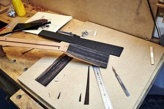 A Richlite fingerboard sits alongside a guitar neck being fitted with an ebony veneer, in a build that mixes natural and synthetic materials.