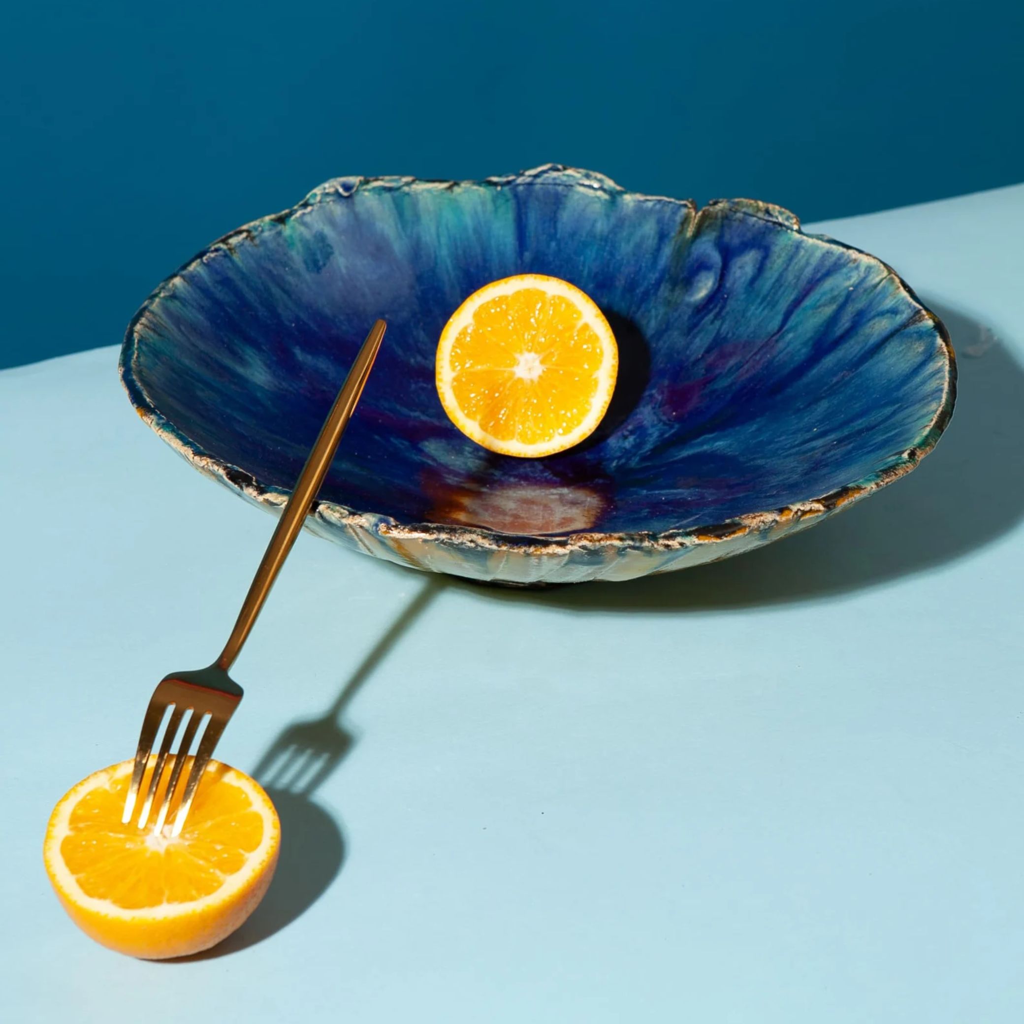 SagaPottery Sculptural Ceramic Platter in blue, set against a pale blue background and filled with half a lemon, the other half of which sits with a fork in its middle next to it.