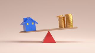 model of blue house and coins balanced on wooden plank on top of red triangle, with blue house side lower than coins