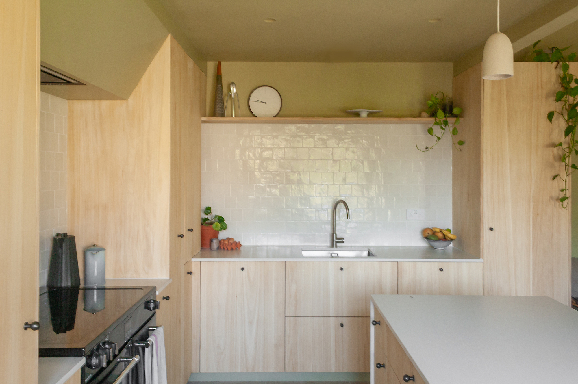 A view of a modern, minimalist kitchen with light wood cupboards and marble worktops, and sage green walls. Above the island is a pendant light and a cooker to the right.