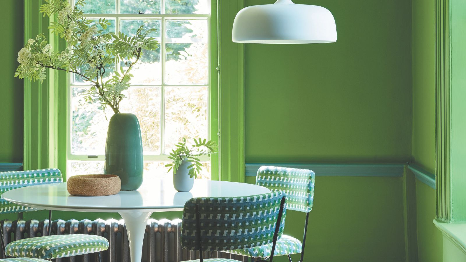 A bright green painting dining room with a white dining table, surrounded by green chairs. A white pendant hanging from the ceiling. 