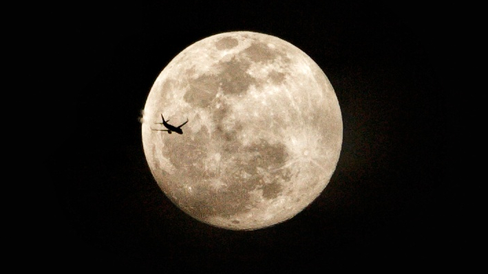 A full moon is pictured against a dark sky with the silhouette of a passenger jet visible to the left of its disk.