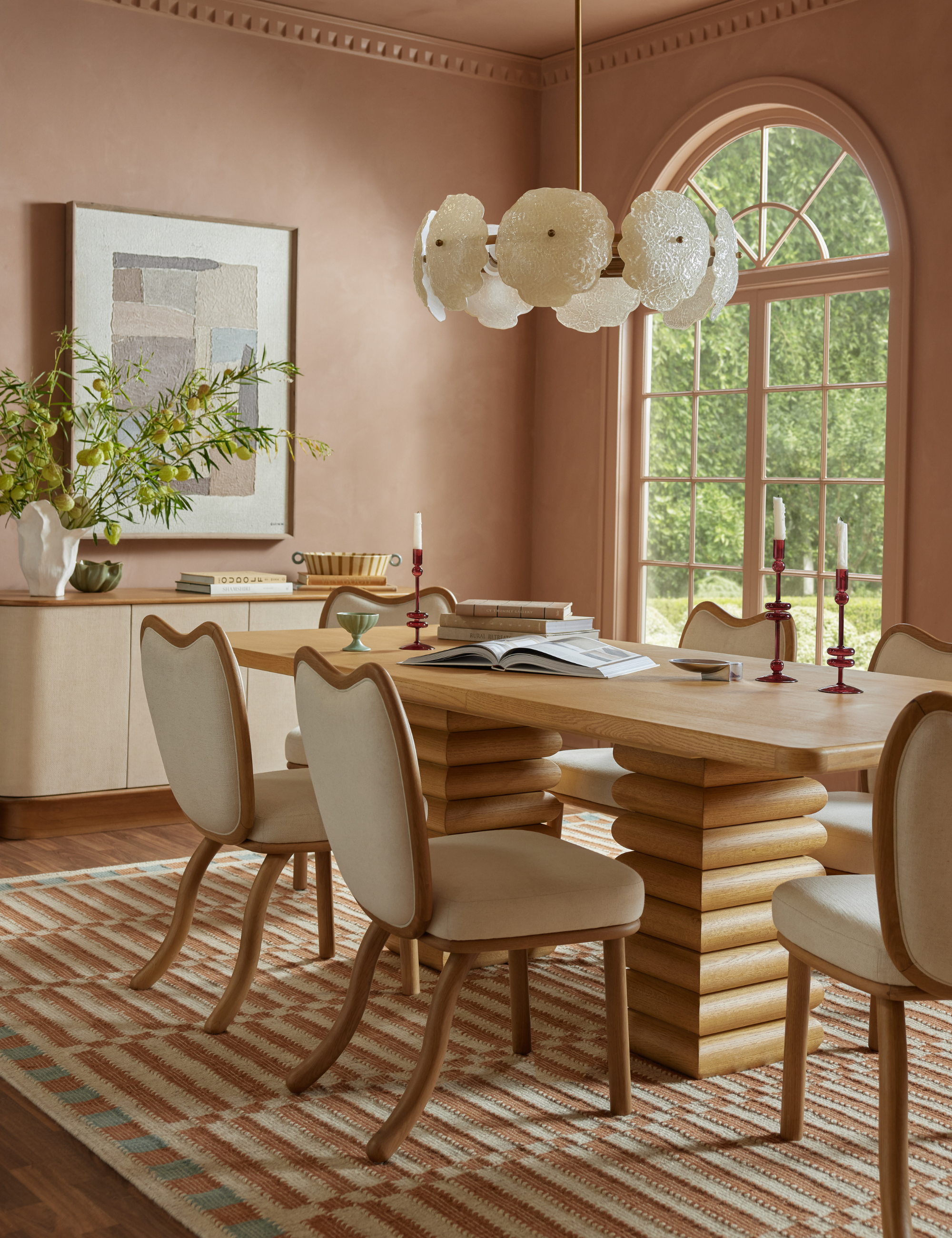 Terracotta-drenched dining room featuring a sculptural wooden double pedestal table accented with playful dining chairs and a floral chandelier overhead.