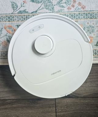 A white robot vacuum on top of dark gray floorboards and a white and green floral rug