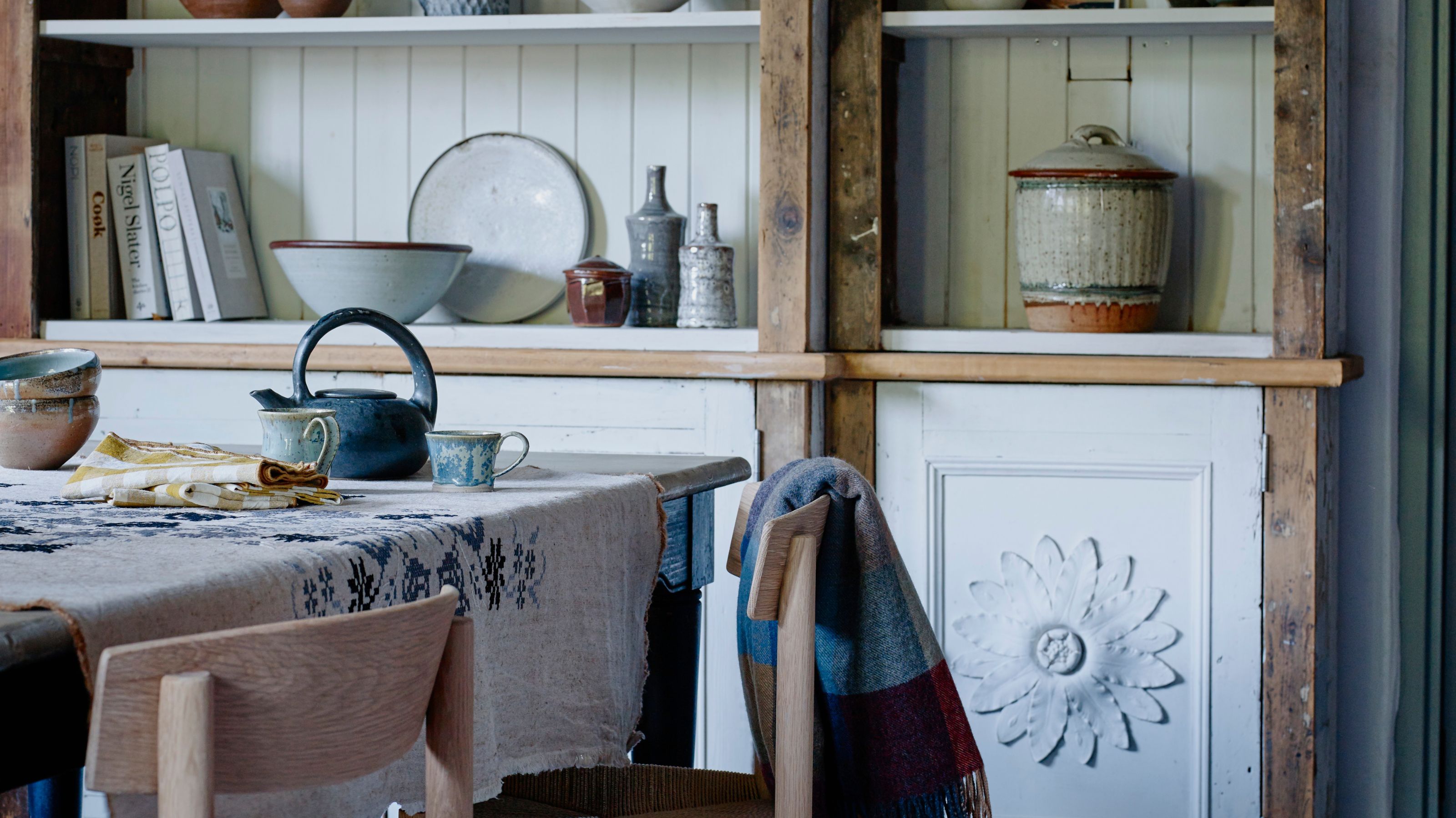 Dining room with dining table with wooden chairs, and shelving with white wall paneling, flower motifs, and assorted ceramics and recipe books