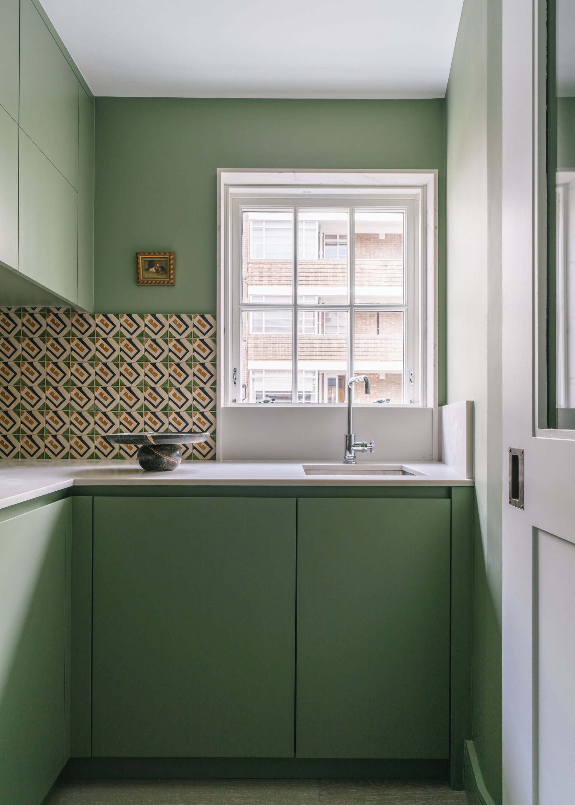 A utility room with sage green cabinets and walls, a small window with a sink in front, and a dark yellow patterned backsplash.