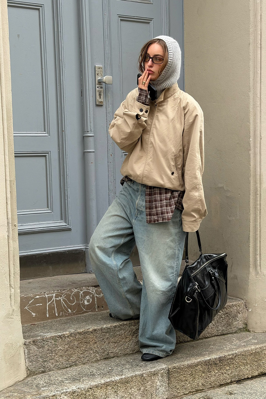 Berlin fashion trends are shown in a street style photo of a woman standing in a doorway wearing a gray balaclava with a tan bomber jacket layered over a dark brown plaid funnel neck shirt styled with ultra-baggy wide leg jeans, black boots, and an oversized black handbag and black eyeglasses