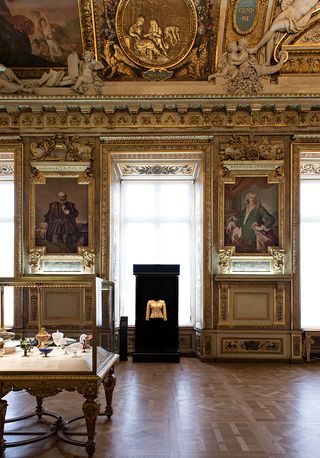 View of The Louvre's tribute to Yves Saint Laurent featuring a black display with a light coloured fashion piece on show in a room with tall windows, wood flooring, paintings, a glass display box with multiple items inside and a decorative ceiling and walls