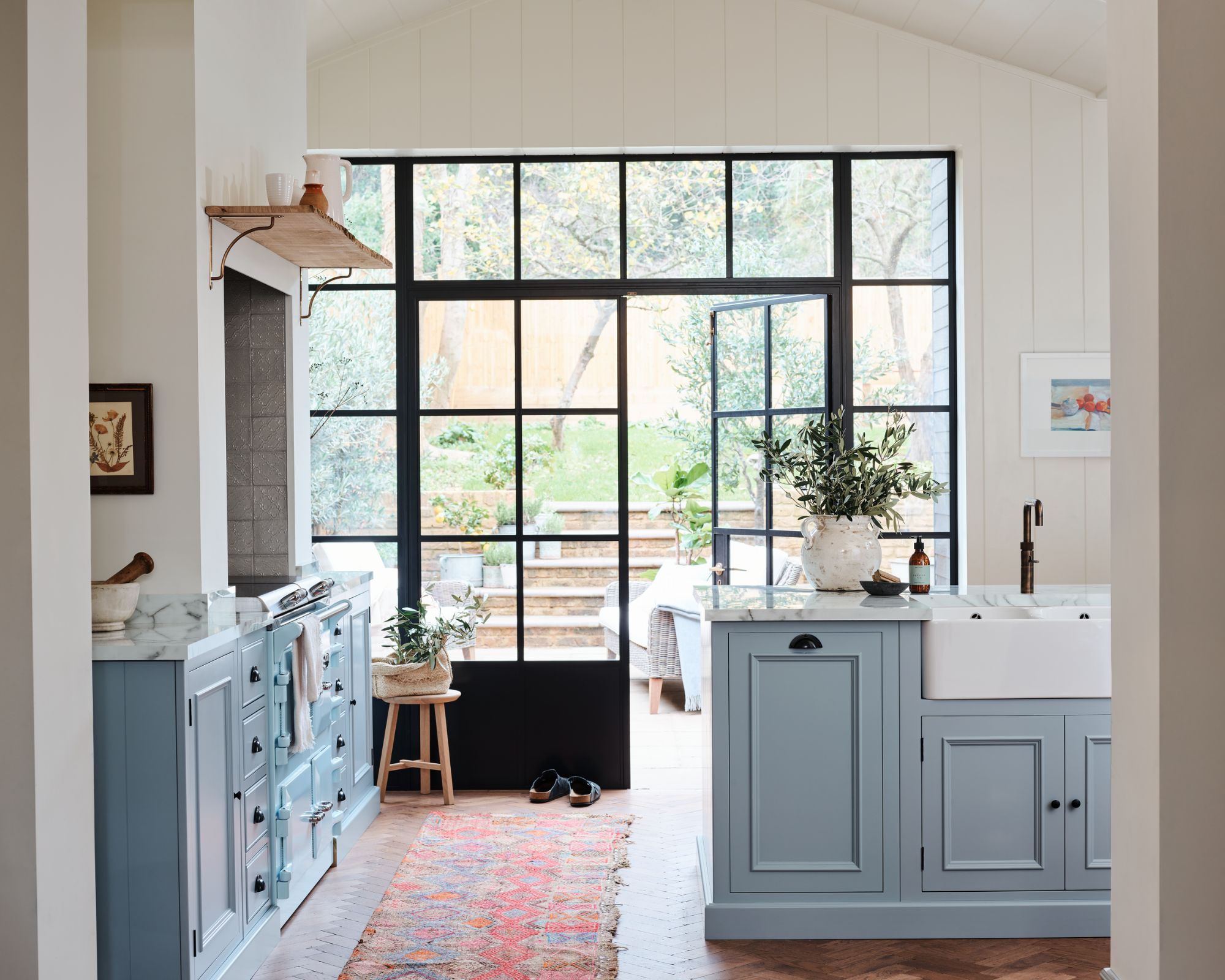 A blue kitchen with large framed windows leading to a garden