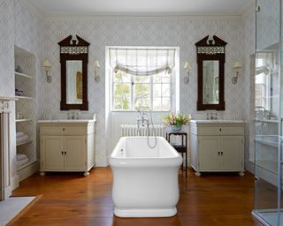 bathroom with patterned wallpaper and freestanding bath, his and hers washstands and wooden floor