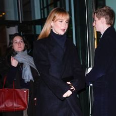 Jennifer Lawrence steps outside wearing a black coat with navy cigarette trousers with a black coat and black asymmetric heels.