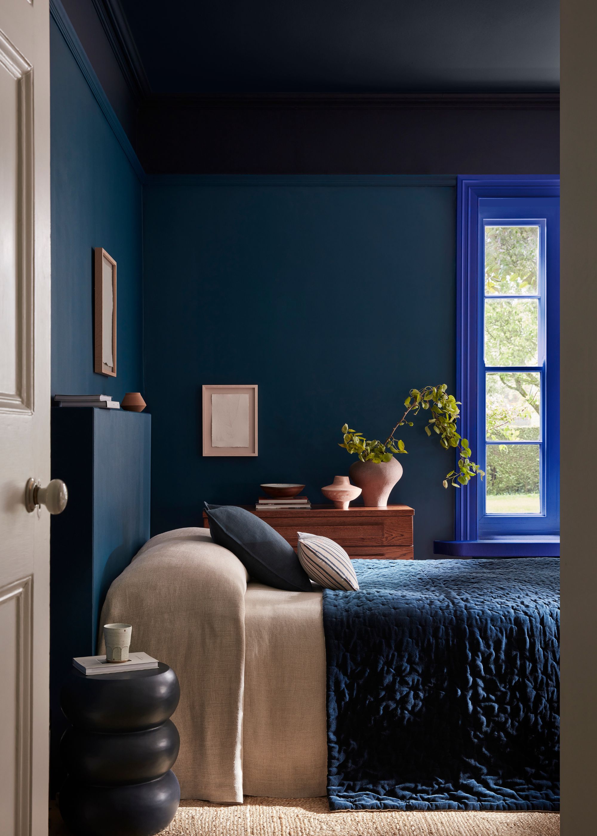 A dark blue bedroom with an electric blue window frame, a dark blue bedcover, and a navy blue nightstand.