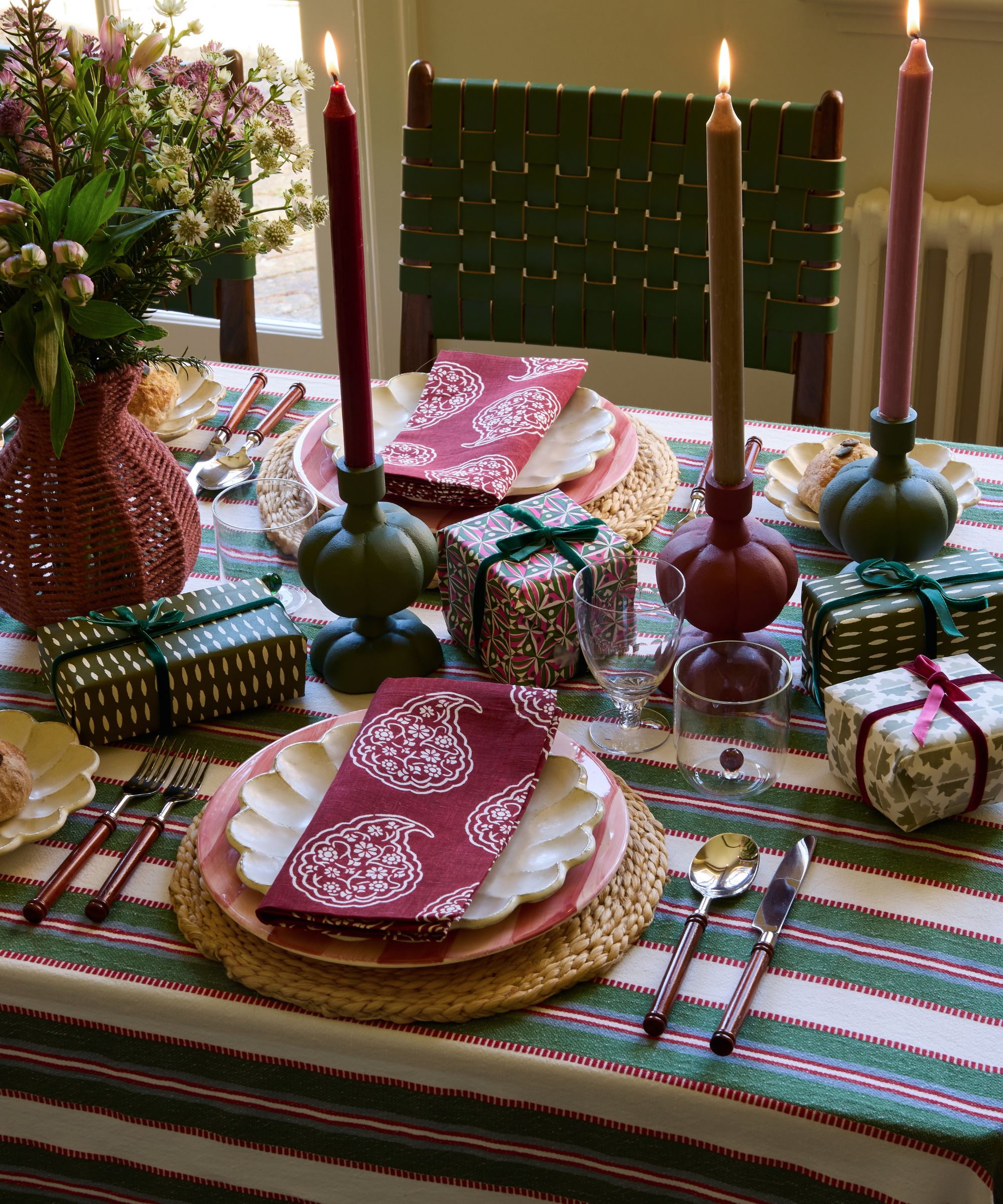 A Christmas tablescape with a green striped tablecloth, layered placemats and plates with burgundy patterned napkins, small wrapped gifts on the table and long dinner candles in candle holders.