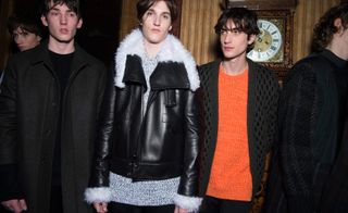Three male models wearing looks from Pringle of Scotland's collection. One model is wearing a black top and coat. Next to him is a model wearing a grey jumper and black leather jacket with white fur. And the third model is wearing an orange jumper and grey and black patterned cardigan