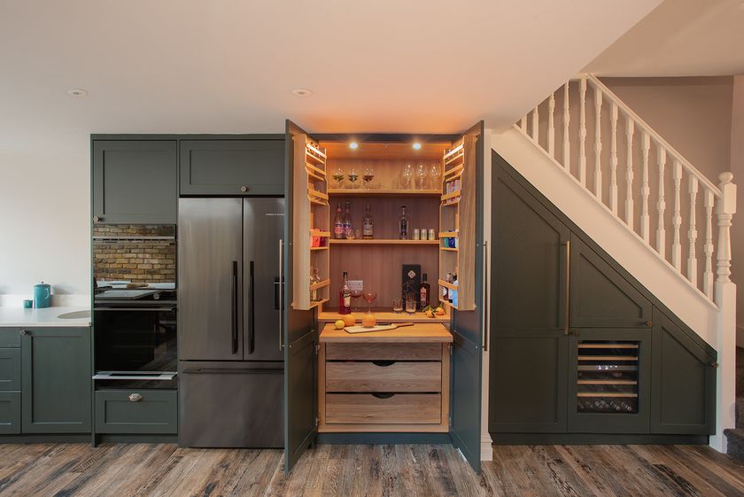 A modern kitchen with dark green cabinetry featuring a built-in drinks cabinet. The open double doors reveal illuminated shelves with glasses, bottles and cocktail ingredients, alongside wooden drawers and integrated under-stairs storage with a wine cooler.