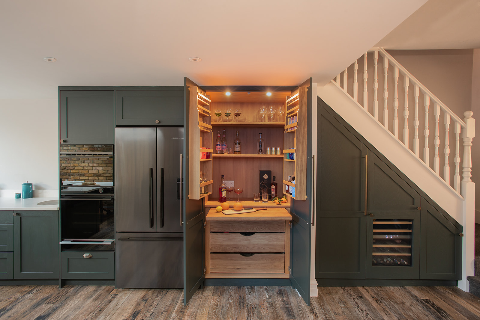A modern kitchen with dark green cabinetry featuring a built-in drinks cabinet. The open double doors reveal illuminated shelves with glasses, bottles and cocktail ingredients, alongside wooden drawers and integrated under-stairs storage with a wine cooler.