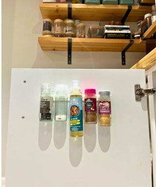 White snack rack inside a white kitchen cabinet that is open, showing left to write pepper jar, salt jar, coconut oil spray, mixed herbs, and garlic granules. Open shelving and glass canisters are visible in the background