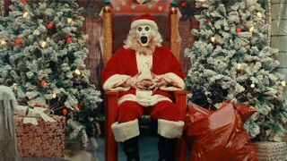 Art the Clown (David Howard Thornton) dressed as Santa Claus sitting on a throne flanked by Christmas trees and sacks of presents in "Terrifier 3"