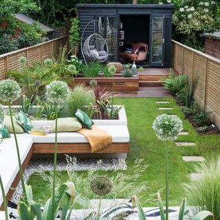 Smartly landscaped narrow garden with raised seating and planters breaking up a green lawn and a path leading to a black garden building