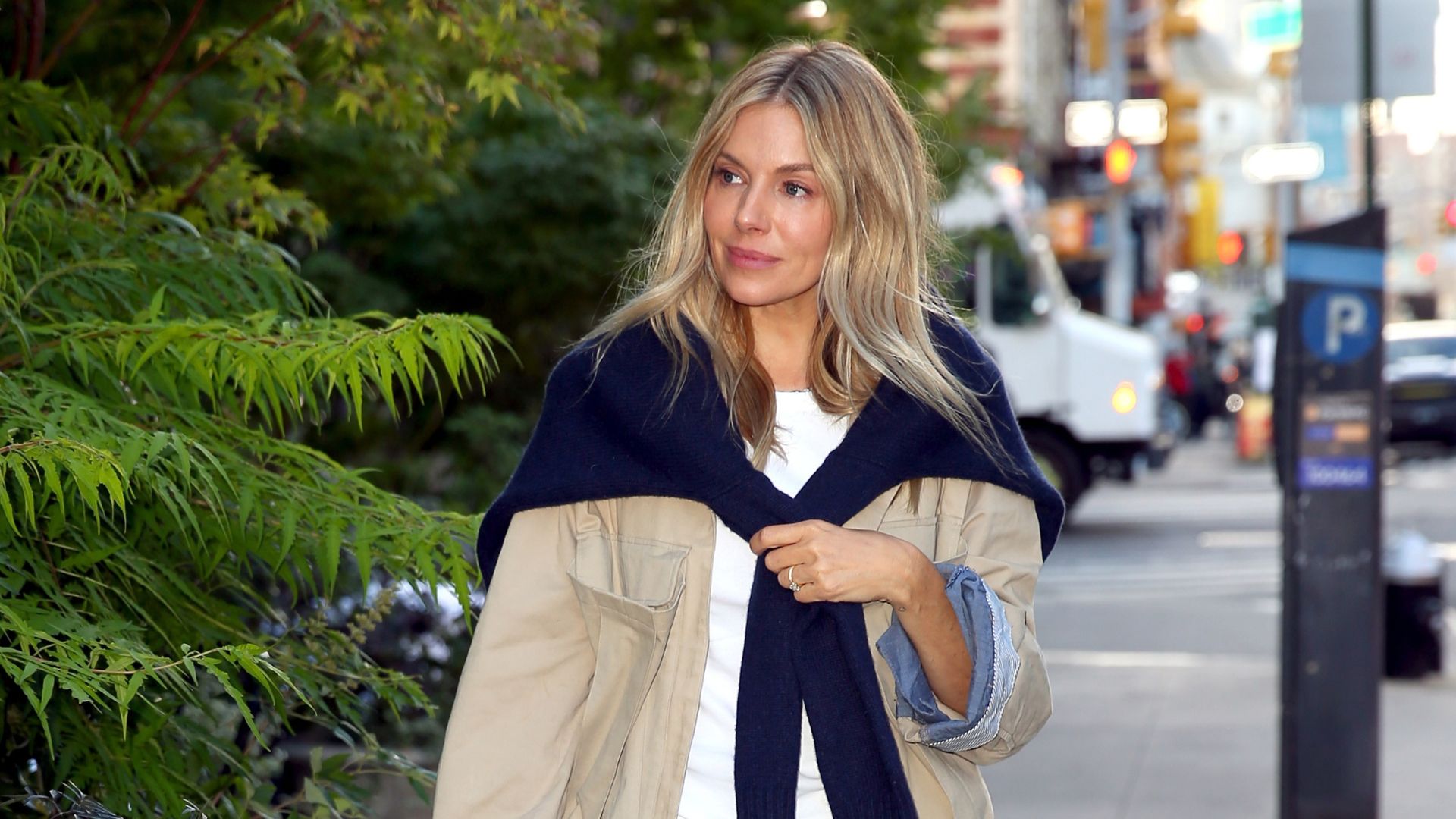 An image of Sienna Miller wearing a white t-shirt, blue jeans, red shoes, beige jacket and navy knit whilst out in New York. 