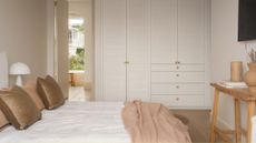 A bright bedroom with a white wardrobe, a wood stool, a white bed woth peach and brown pillows, and a mushroom lamp