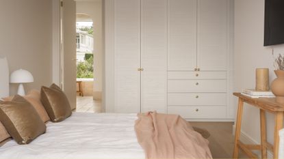 A bright bedroom with a white wardrobe, a wood stool, a white bed woth peach and brown pillows, and a mushroom lamp