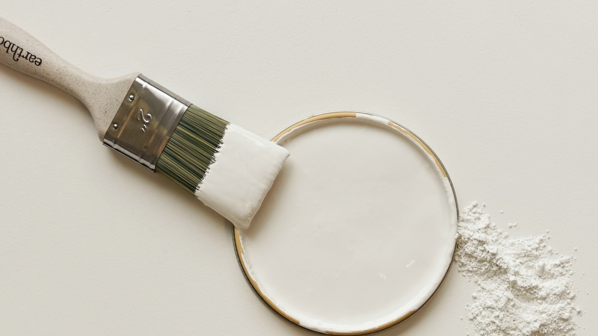 Paintbrush dipped in white paint beside an open tin, with a scattering of loose plaster or powder.