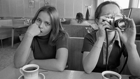 Two young women sit in a diner booth with cups of coffee, one resting her chin on her fist and staring at the camera while the other points a 35mm camera straight back at the photographer.