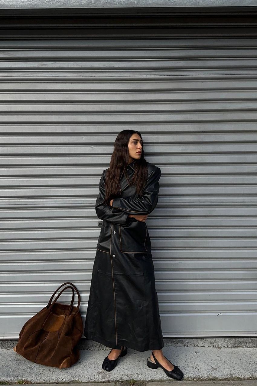 A woman standing outside wearing a long leather jacket as a dress with black ballet flats and a brown suede tote bag.