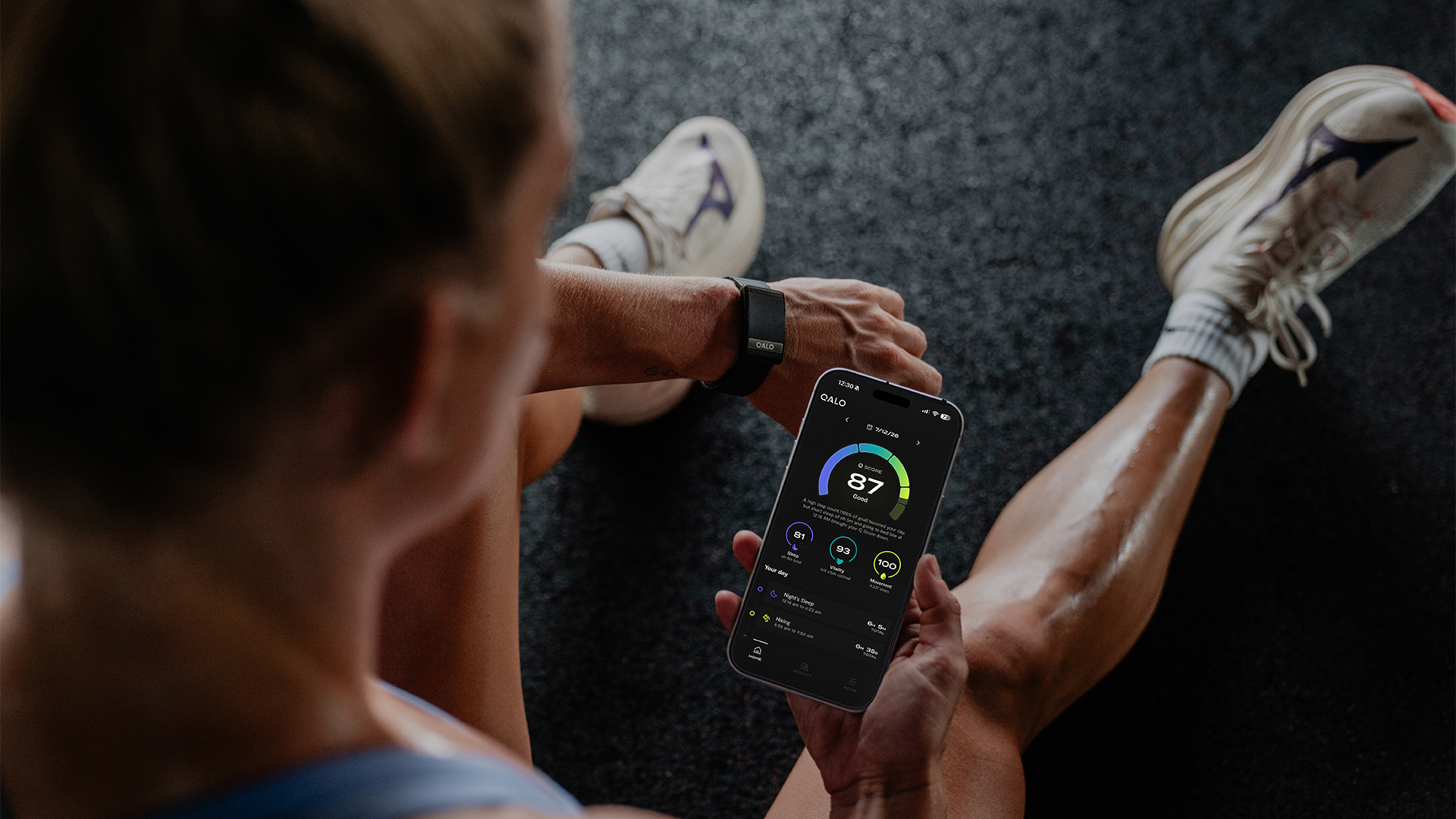 A close up of a person sitting on the floor, wearing a QALO SL-1 smart band and checking the QALO app for their Q score