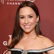 Lacey Chabert smiles while wearing an off-the-shoulder black dress with a silver ribbon trim