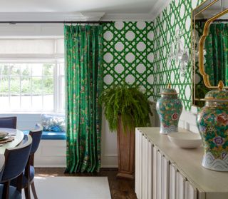 dining room with green trellis wallpaper and green chinoiserie curtains with blue window seat cushion and dining chairs and white ridged console with green ginger jars and oval mirror above