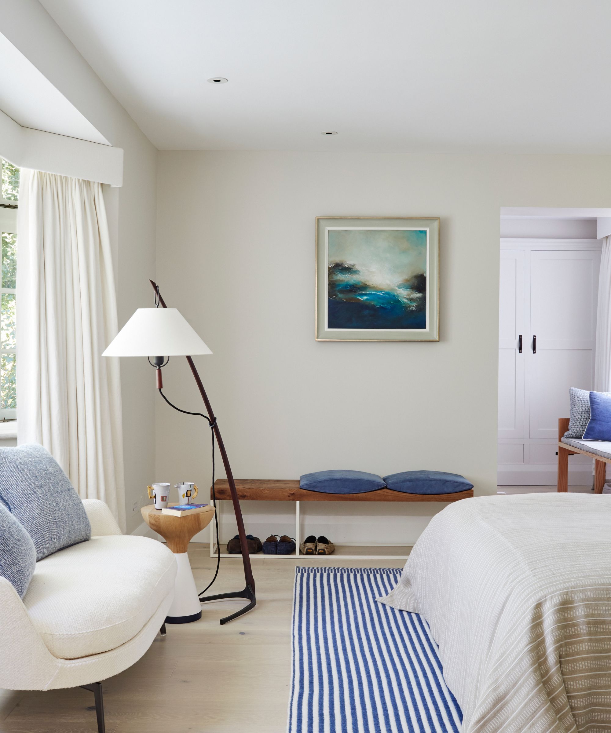 A bright bedroom with white and blue accents, a white boucle sofa, a white and blue striped rug, a wooden bench, and a framed painted seascape.