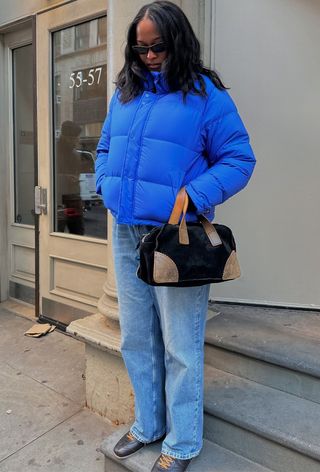 a roundup of the best puffer jackets shown in a photo of a woman wearing a bright blue puffer coat with baggy jeans, a black suede bowler bag, and black leather sneakers