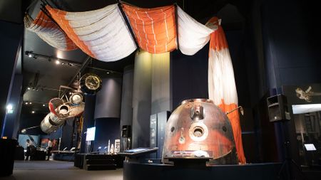 a space capsule and its orange and white parachute are seen on display with other space artifacts in a museum