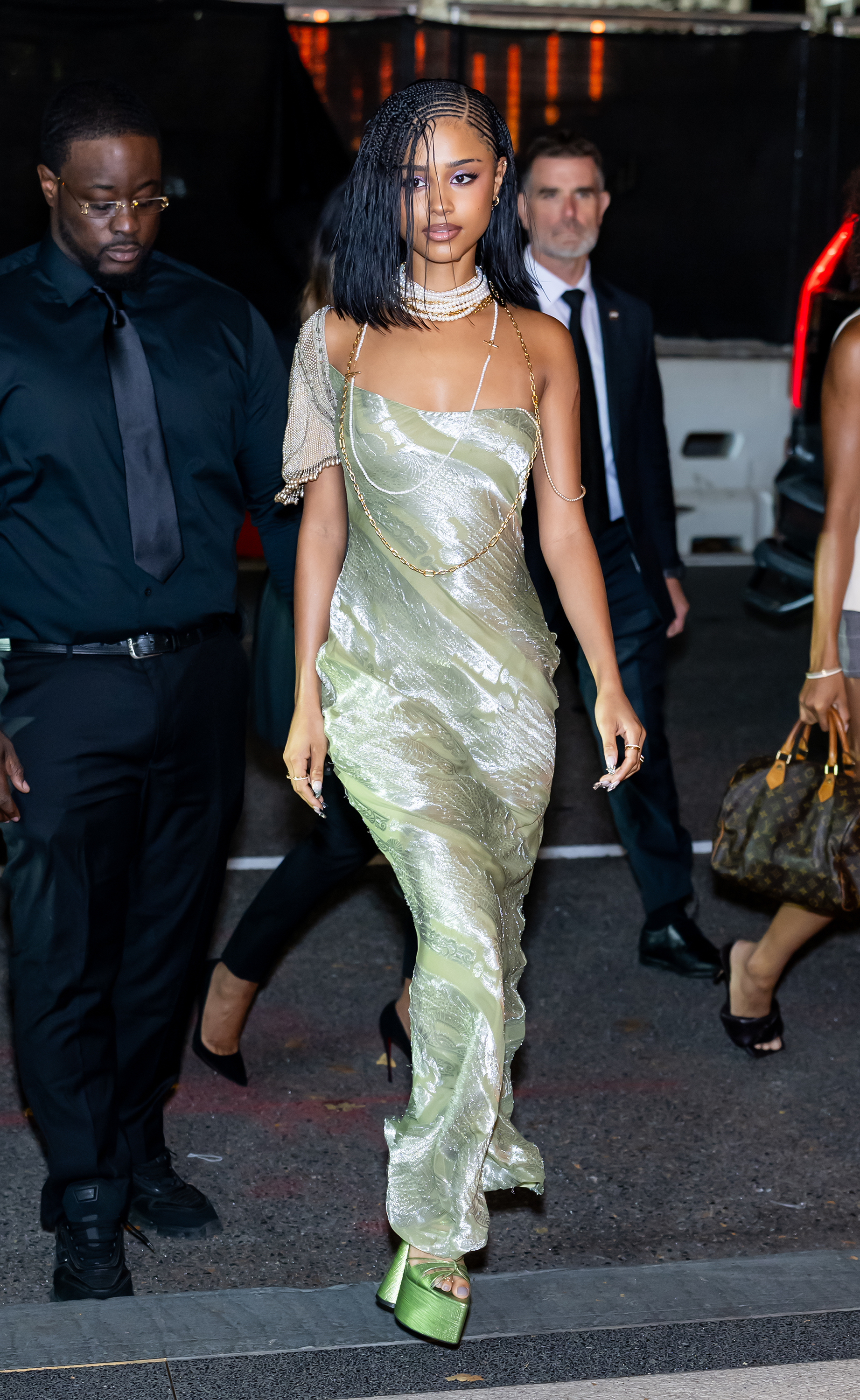 Tyla wearing a braided bob hairstyle with cornrows in the front. She is wearing a green silk dress with lace detailing woven throughout the dress and green platform heels