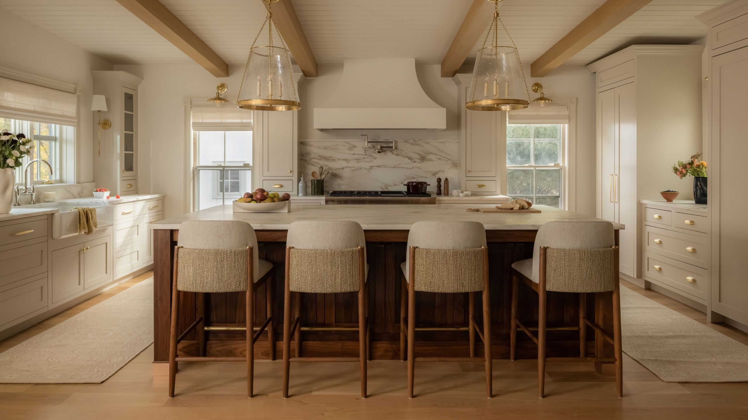 a large, warm beige kitchen with ceiling beams and panels, a dark wooden island with woven stools and glass pendants overhead, range cooker with plaster hood and marble counters