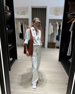 @elizagracehuber taking a mirror selfie wearing white button-down, white pants, and tan bag