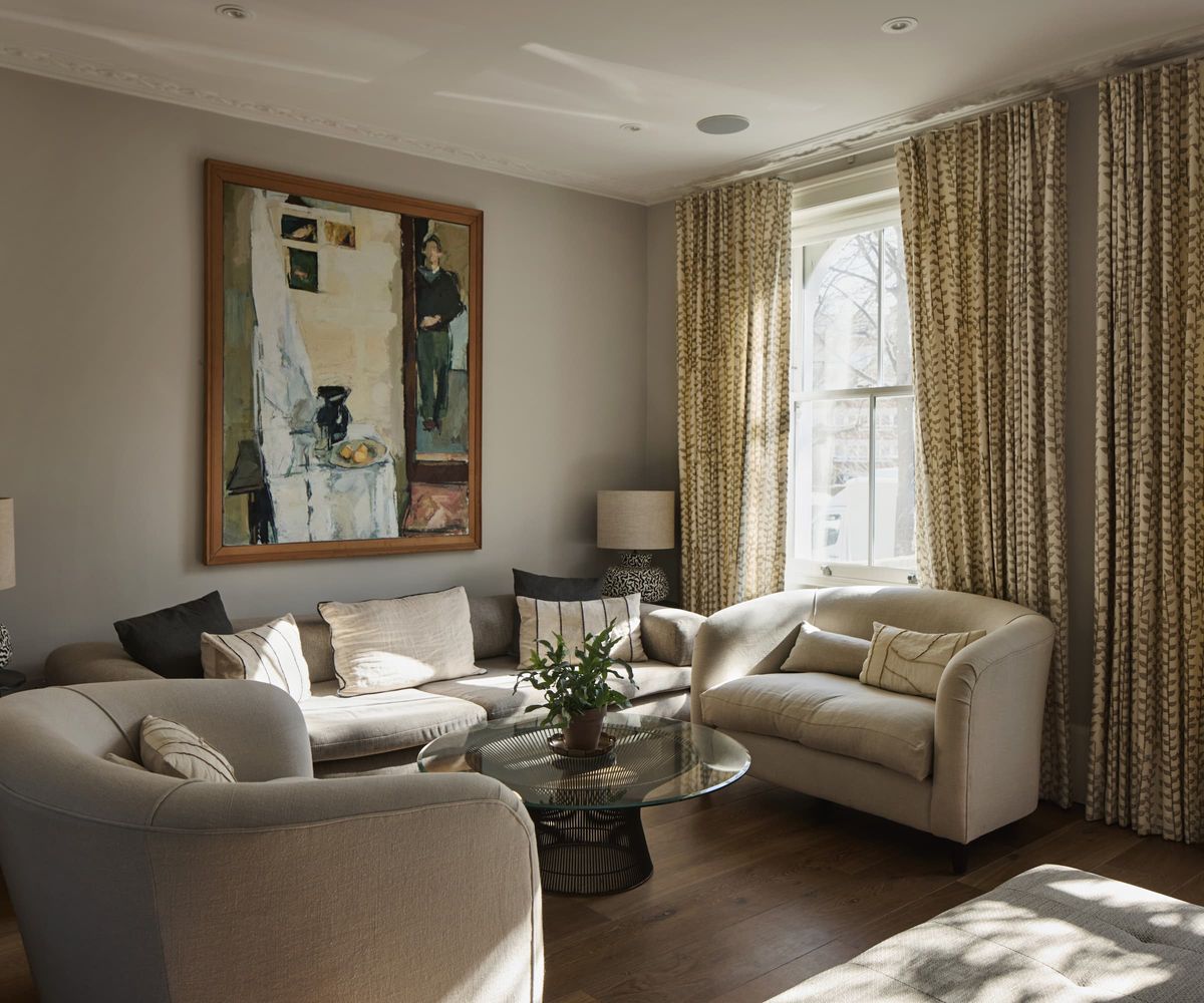 A living room with large curtains and three white sofas and a picture on the wall