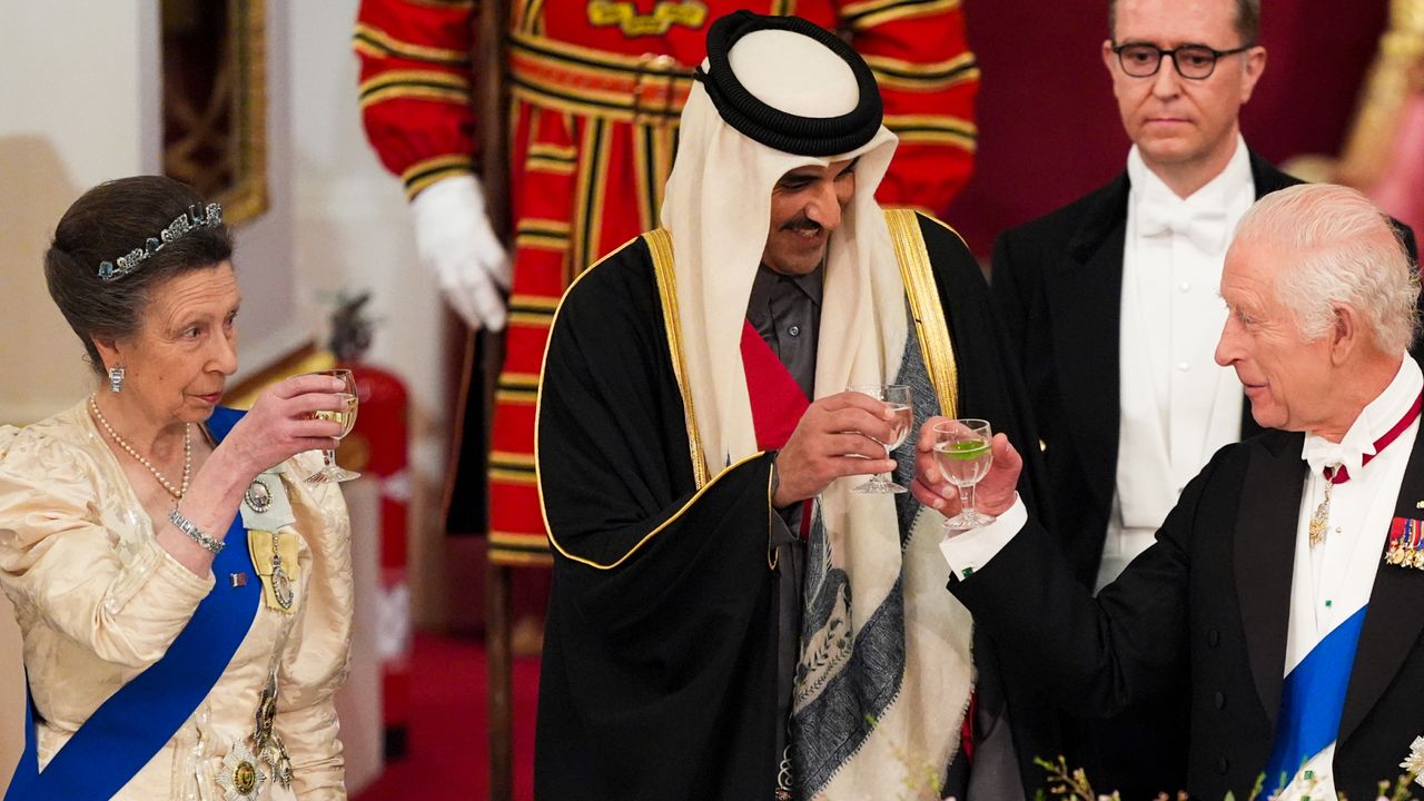 Princess Anne holds up a glass while wearing a cream dress and a small tiara