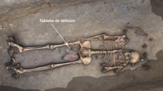 A skeleton is lying in the ground with its skull at the right. There are artifacts between the legs near the pelvis, with an arrow pointing to a "tablette de defixion" or curse tablet.