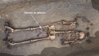 A skeleton is lying in the ground with its skull at the right. There are artifacts between the legs near the pelvis, with an arrow pointing to a "tablette de defixion" or curse tablet.