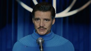 Sporting a blue pullover, Pedro Pascal, as Mister Fantastic, addresses a microphone in his Fantastic Four: First Steps role as Reed Richards.