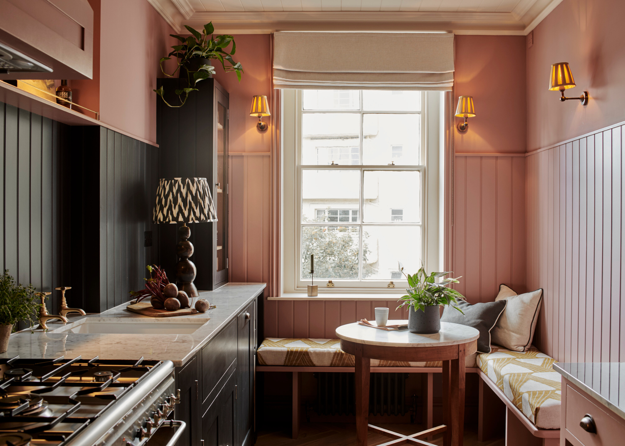 A small pink and black kitchen with matchboard panelling on the walls, banquette seating beneath the window and around the corner and warm wall lamps above this