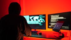 Hacker working on a desktop computer with dual monitors in a room with red lights.