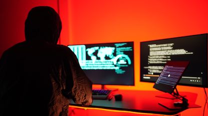 Hacker working on a desktop computer with dual monitors in a room with red lights.