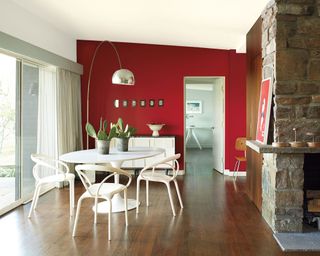 Large dining room with red painted feature wall, dark wooden flooring, white tulip dining table with white chairs, white sideboard, metallic twiggy table lamp over table, traditional brick fireplace, dining table beside large sliding French doors