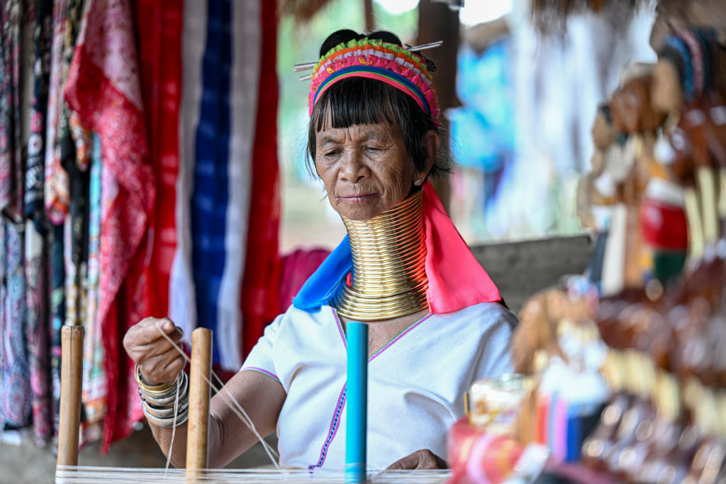 A woman wears a series of gold neck rings to lengthen her neck. She also wears a pink scarf in her hair and a white shirt and sits in a well decorated tent