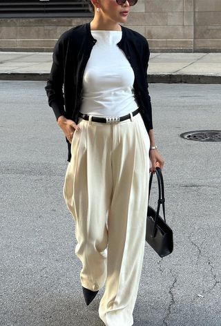 a photo showing what to wear to work with a woman wearing white pleated trousers with a white top, black cardigan, black belt, black pumps, and a black shoulder bag